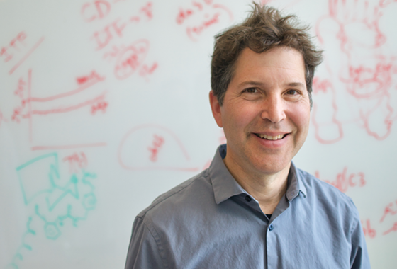 David Baker smiling at the camera in front of a whiteboard with equations and drawings